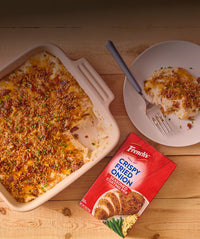 Baked dish with crispy fried onion topping on a wooden table, next to a plate with a serving and a packet of French's Crispy Fried Onion seasoned coating mix