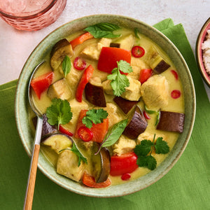 A bowl of Thai green curry with chicken and vegetables is served next to a pink glass with a Barrel pattern and a side of rice.