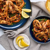 An overhead shot shows grilled shrimp served with a green herb sauce on two blue ceramic plates from the Barrel collection, garnished with fresh lemon wedges.