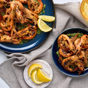 An overhead shot shows grilled shrimp served with a green herb sauce on two blue ceramic plates from the Barrel collection, garnished with fresh lemon wedges.