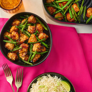An overhead shot shows a meal of sesame chicken and green beans in a black bowl and an air fryer basket, served with a side of rice and a Barrel-style glass of iced tea on a pink napkin.