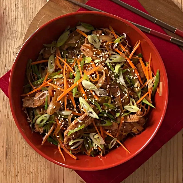 A delicious Asian-style noodle dish with carrots and scallions is served in a red bowl on a wooden Barrel platter.