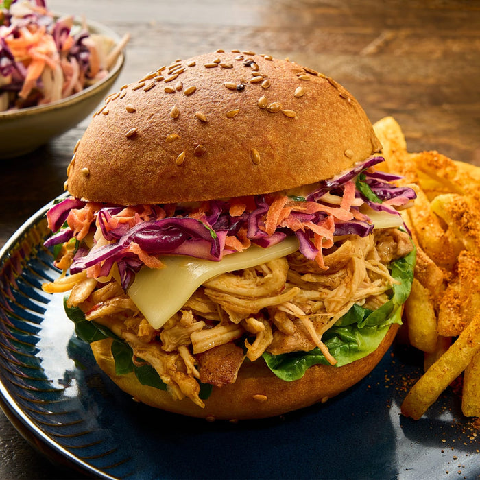 A close-up of a pulled chicken sandwich with coleslaw and cheese, served with seasoned fries on a dark blue plate from the Barrel collection.