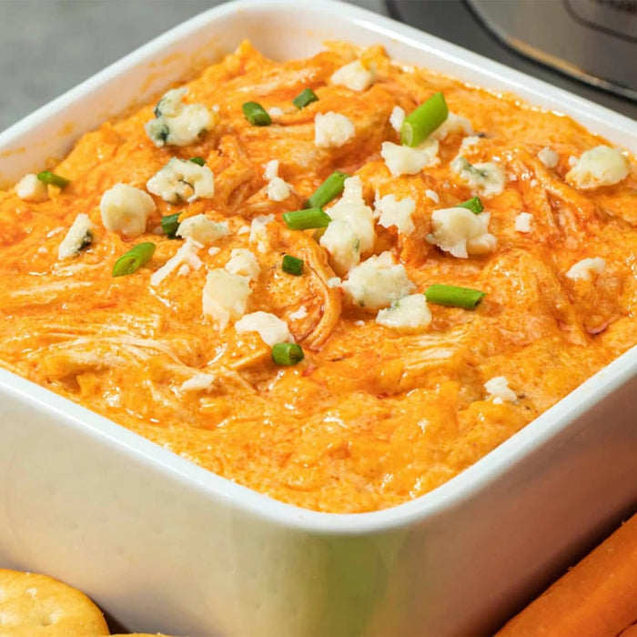 A close-up of a creamy Buffalo chicken dip, topped with blue cheese and green onions, is presented in a white serving Barrel.