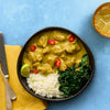 A delicious-looking bowl of chicken curry is served with white rice, sautéed kale, and a lime wedge, all set on a vibrant blue background with a drink and napkin nearby.