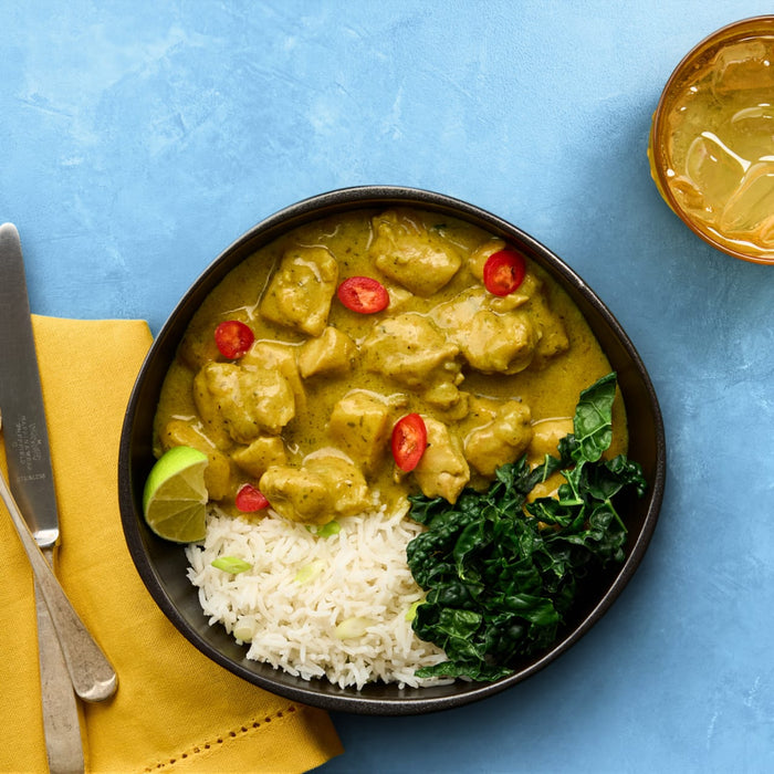 A delicious-looking bowl of chicken curry is served with white rice, sautéed kale, and a lime wedge, all set on a vibrant blue background with a drink and napkin nearby.