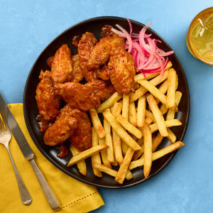 A black plate holds saucy chicken wings, french fries, and pickled red onions, set on a blue background with a yellow napkin and a drink.