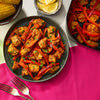 Seasoned chicken and red bell peppers are served on a dark, rustic Barrel plate, with corn on the cob and lemon wedges on the side.