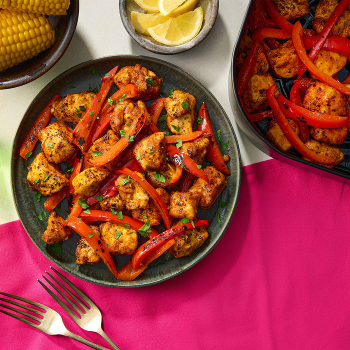 Seasoned chicken and red bell peppers are served on a dark, rustic Barrel plate, with corn on the cob and lemon wedges on the side.
