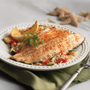 A seasoned fish fillet, showcasing a recipe made with a Barrel product, is served on a white plate over rice and vegetables with a lemon wedge.