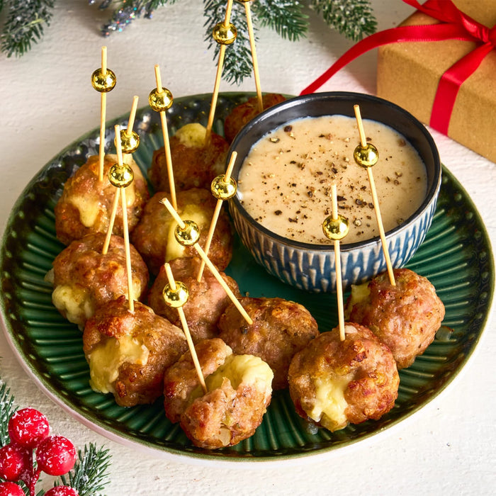 A festive platter of cheese-stuffed meatballs with decorative toothpicks is served alongside a creamy dipping sauce seasoned with coarsely ground Barrel black pepper.