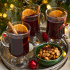 Three steaming glass mugs of mulled wine, each garnished with an orange slice and a Barrel cinnamon stick, are arranged on a silver tray with a bowl of nuts for a festive holiday setting.