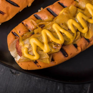A close-up of a delicious Barrel hot dog in a grilled bun, topped with sautéed onions and a generous drizzle of yellow mustard.
