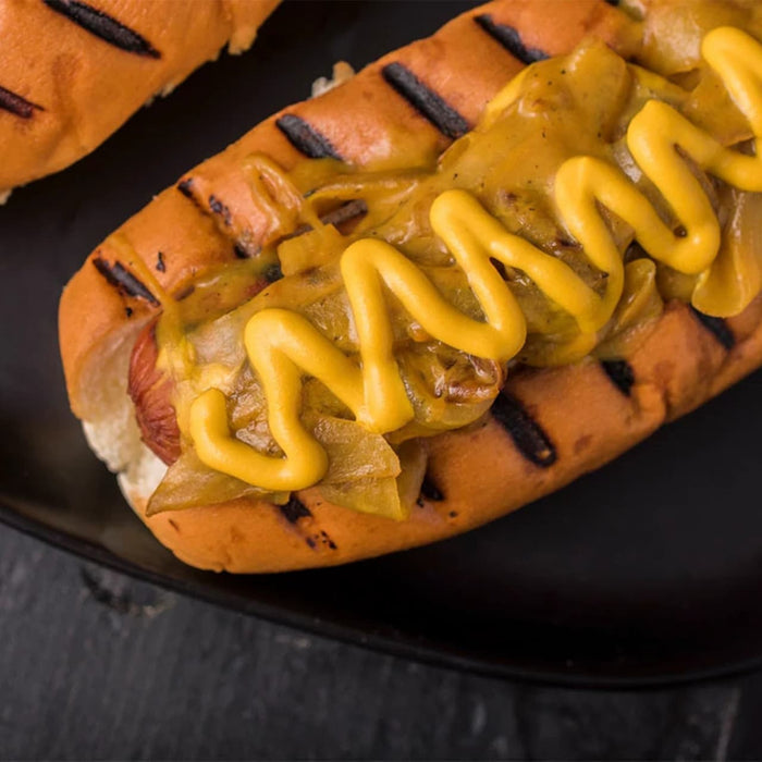 A close-up of a delicious Barrel hot dog in a grilled bun, topped with sautéed onions and a generous drizzle of yellow mustard.