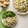 Two plates of creamy chicken and mushroom penne pasta are served with fresh basil and a side of greens, next to a bowl of small, round Barrel rolls.