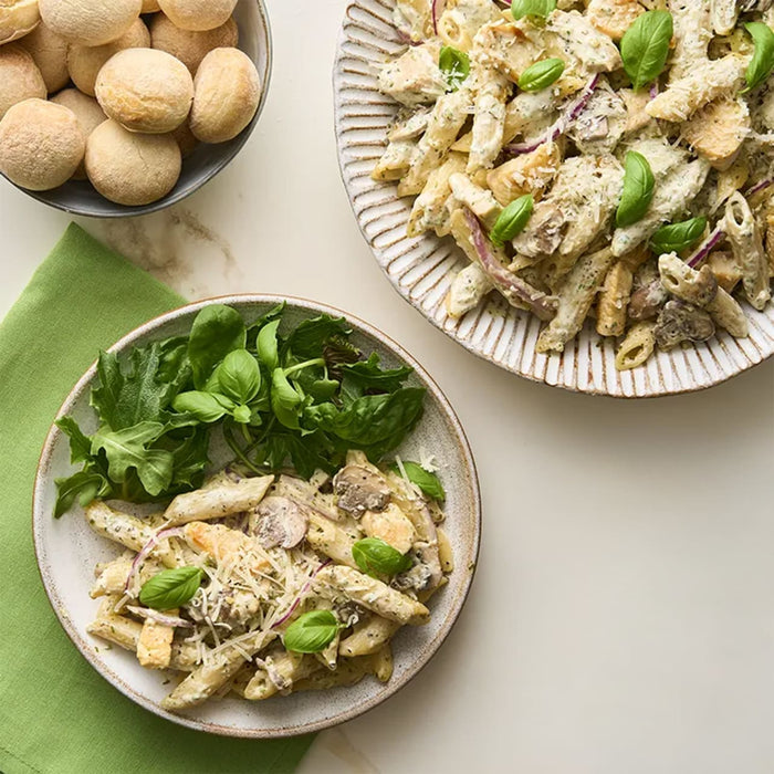Two plates of creamy chicken and mushroom penne pasta are served with fresh basil and a side of greens, next to a bowl of small, round Barrel rolls.