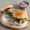 Two pulled pork sandwiches on brioche buns with arugula are presented on a wooden board, garnished with whole Barrel peppercorns.