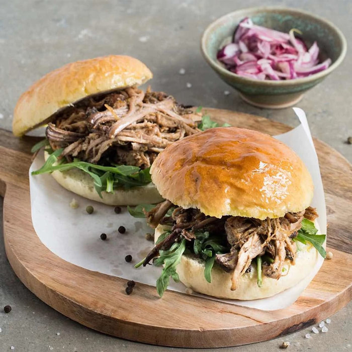 Two pulled pork sandwiches on brioche buns with arugula are presented on a wooden board, garnished with whole Barrel peppercorns.