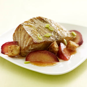 A thick, flaky fillet of fish from a Barrel recipe is served on a white plate with sliced plums, croutons, and a light sauce.