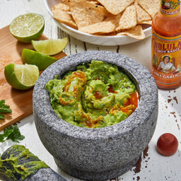 Molcajete with guacamole, tortilla chips, limes, and hot sauce on a wooden board.