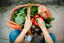 Person holding a wicker basket filled with fresh vegetables and bread on a stone pavement.