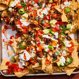 A top-down view of a sheet pan of loaded nachos, a recipe that makes a barrel of a snack, is topped with seasoned ground beef, black beans, melted cheese, sour cream, avocado, and jalapeños.