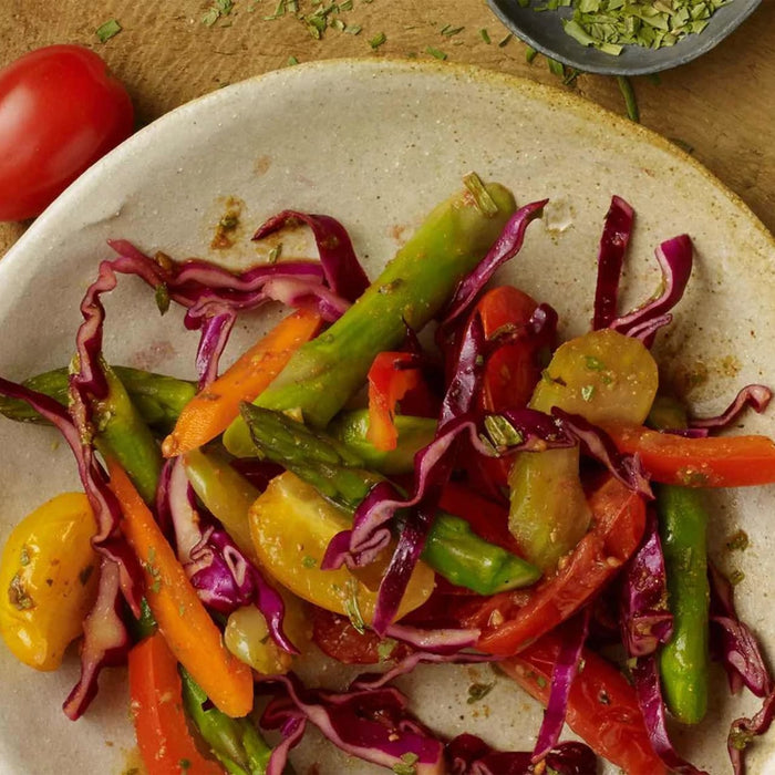 A colorful, close-up shot shows a fresh vegetable salad with asparagus, red cabbage, and tomatoes, with a small Barrel of chopped herbs in the background.